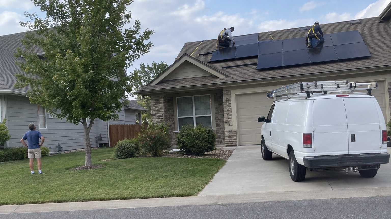 Two installers mounting solar panels on a roof while a neighbor watches from the lawn next door