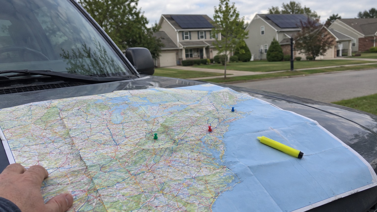 A road map of the eastern United States held flat on a truck hood with push pins in it, and houses with solar panels behind