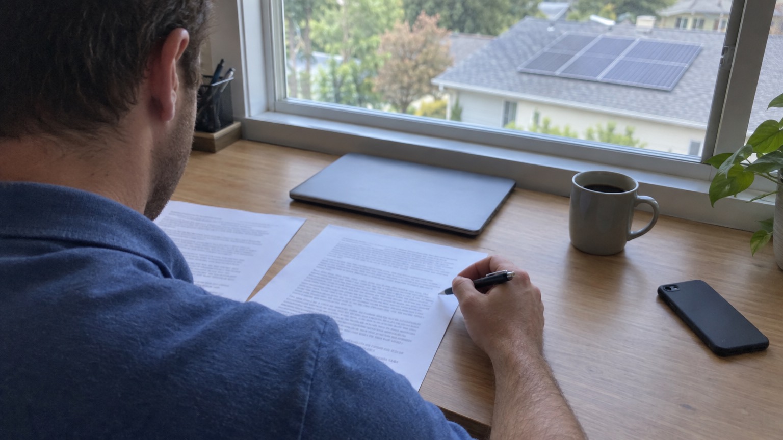 Seen over his shoulder, a man at a home office desk reads two printed proposals with a pen in his hand, a closed laptop beside them